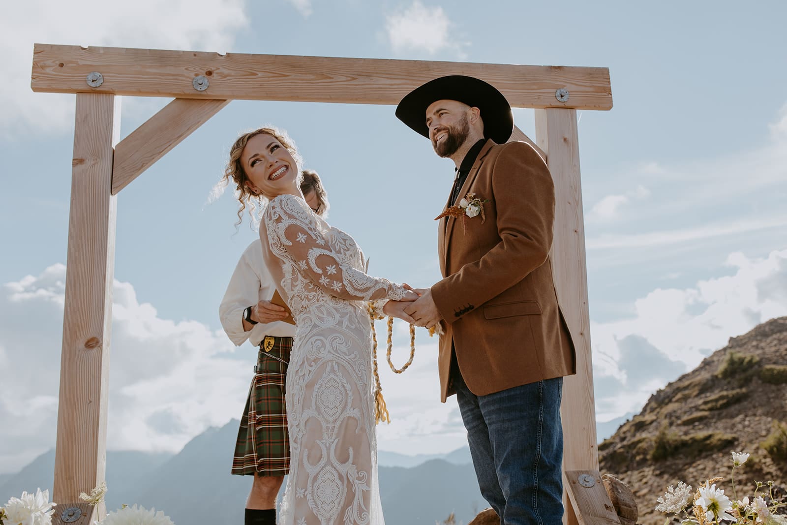 A bride and groom stand , holding hands on their wedding day during their elopement ceremony in the mountains. The bride is looking over her shoulder, smiling.